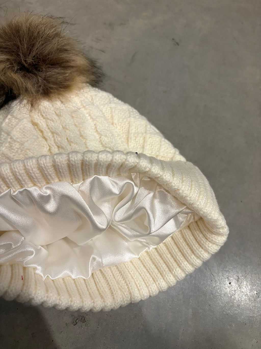 White knit beanie with fur pom-pom on a gray surface showing the satin lining