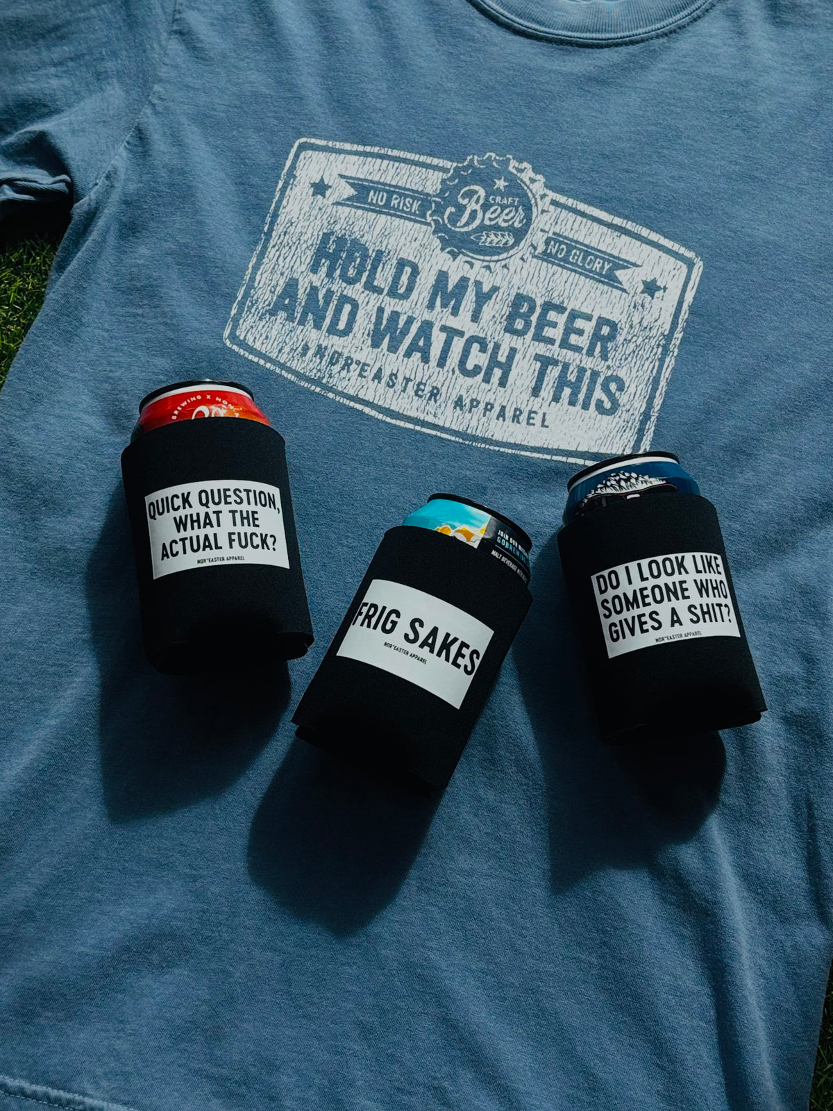 A blue graphic t-shirt laid flat on grass, featuring a large distressed white print that reads 'HOLD MY BEER AND WATCH THIS' with 'No Joke Beer Brand' and 'Independent Apparel' in the crest. Three black can coolers holding beer cans are arranged in front of the shirt.