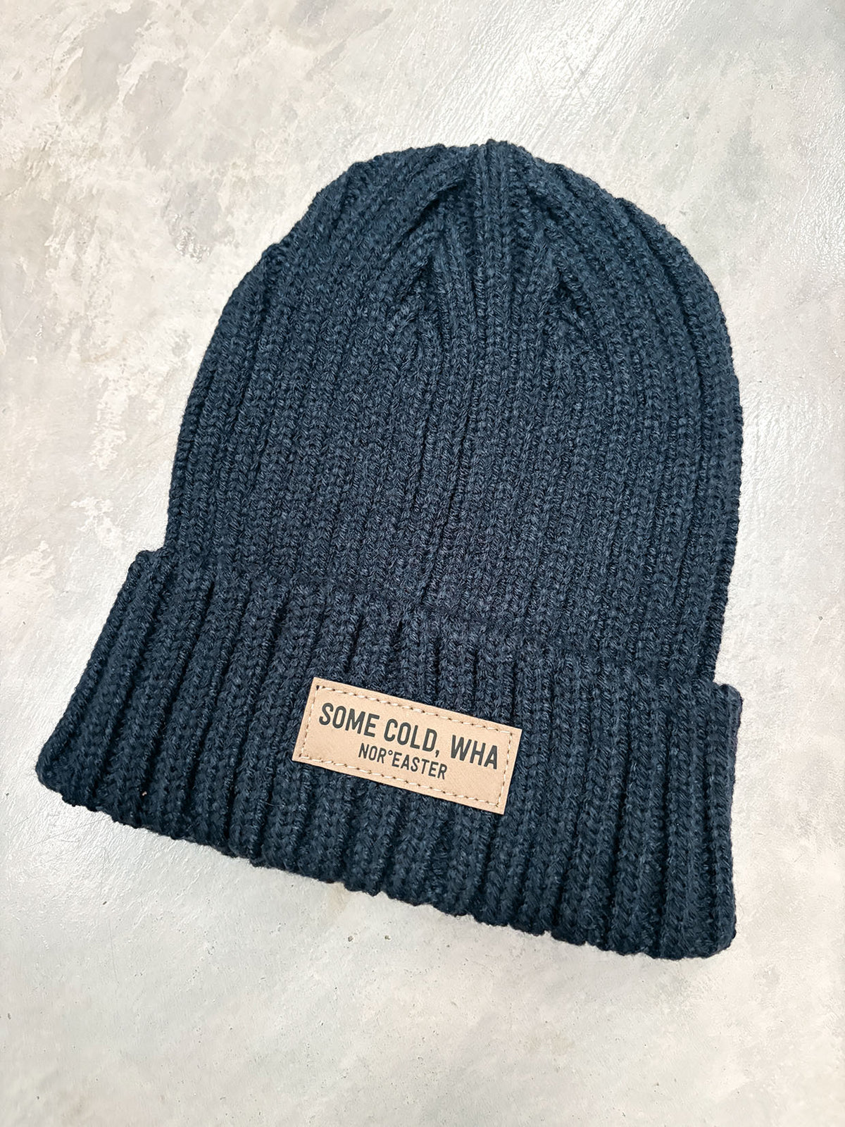 A dark navy blue ribbed knit beanie hat with a small tan rectangular patch on the cuff that reads 'SOME COLD, WHA NON-EASTER'. The hat is photographed from a top-down angle on a light gray textured surface.