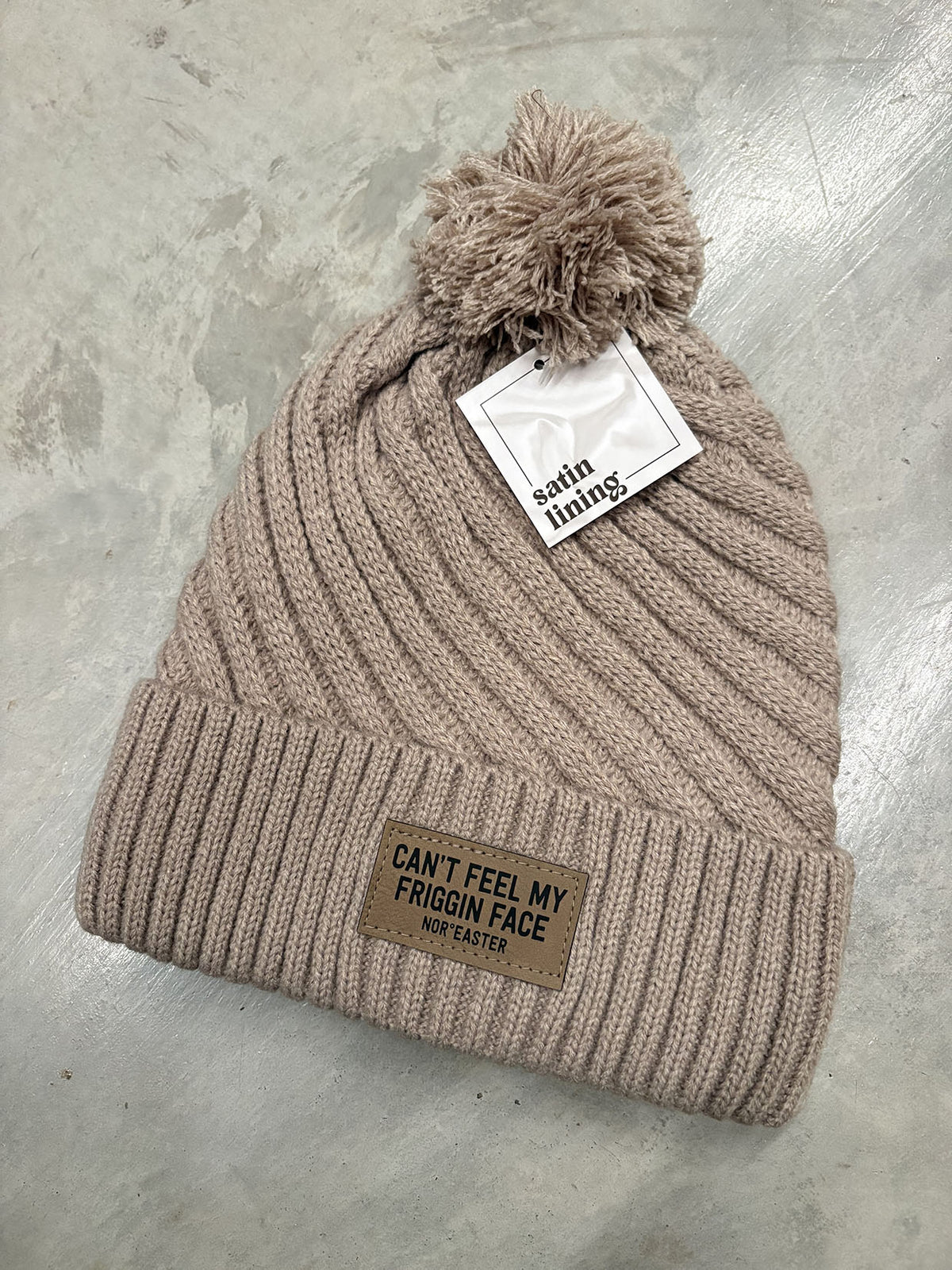 A ribbed knit pom-pom beanie in a warm taupe-beige color, featuring a brown leather patch on the cuff that reads 'CAN'T FEEL MY FRIGGIN FACE' and a white paper tag reading 'satin lining'. The hat is laid flat on a textured gray concrete surface.