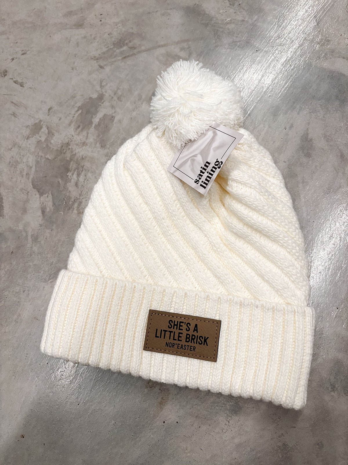 An off-white ribbed knit pom-pom beanie hat with a brown leather patch on the cuff that reads 'SHE'S A LITTLE BRICK' and 'MOR EASTER'. A white paper tag with 'smile things' is attached near the pom-pom.