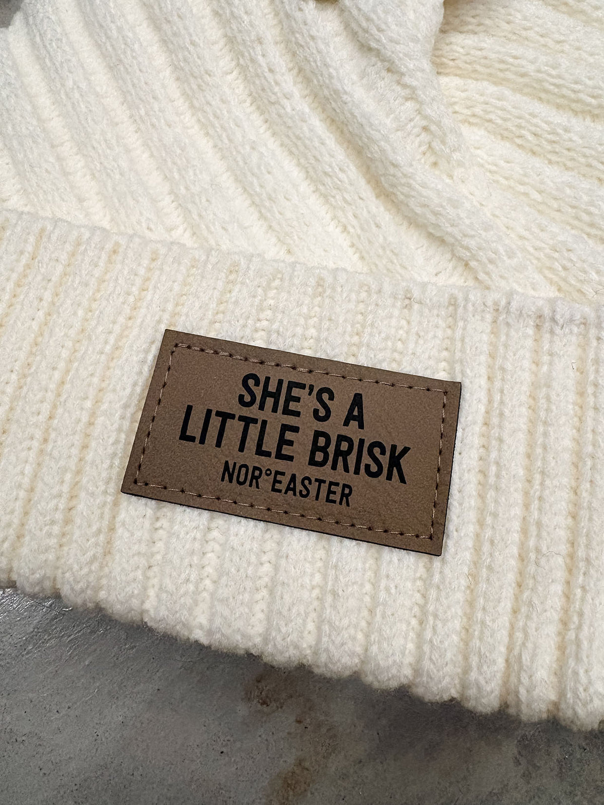 Close-up of a cream-colored ribbed knit beanie with a brown leather patch that reads 'SHE'S A LITTLE BRISK NOR'EASTER'.