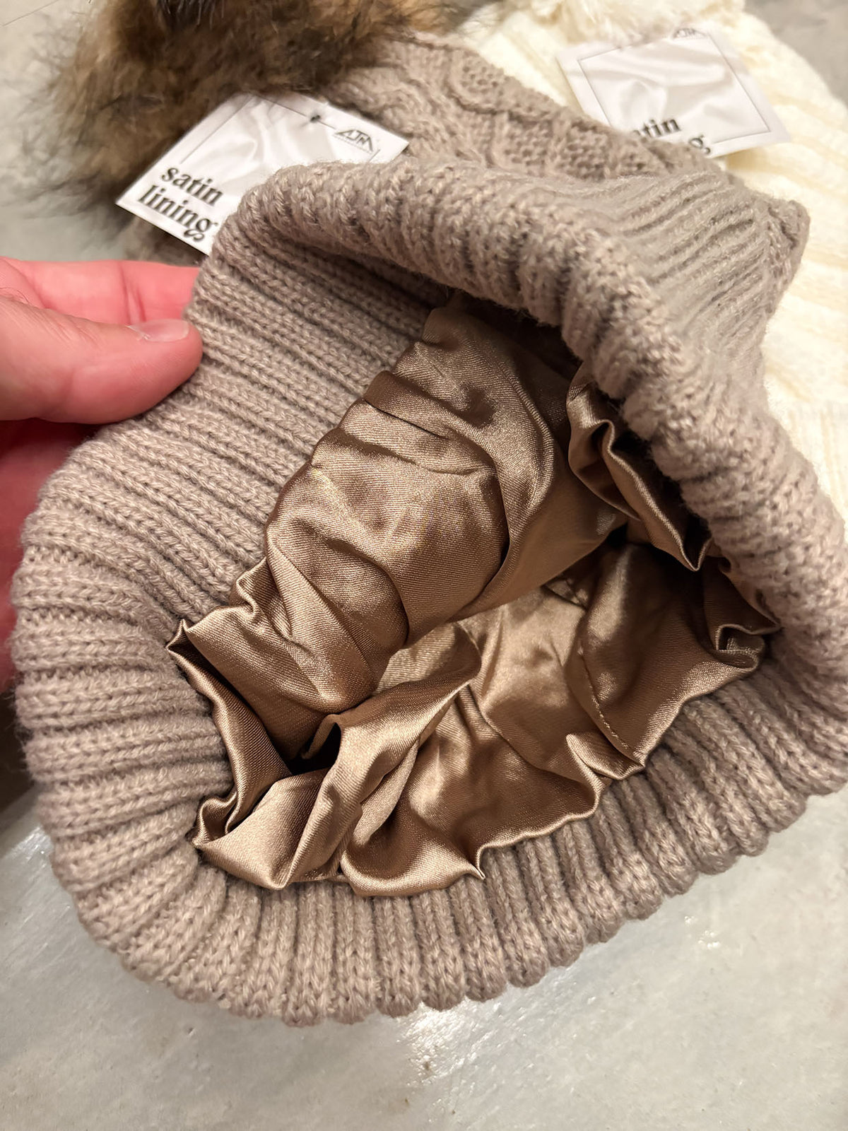 A close-up of a ribbed knit beanie in taupe with a matching satin lining, held open by a hand. The interior features a gathered, ruffled satin lining in a champagne beige color.