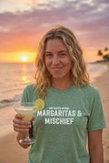 Woman holding a margarita with sunset and beach in the background, wearing a green t-shirt with text.