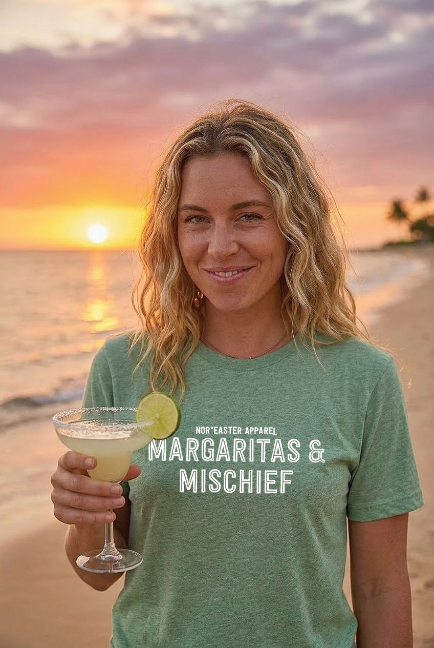 Woman holding a margarita with sunset and beach in the background, wearing a green t-shirt with text.