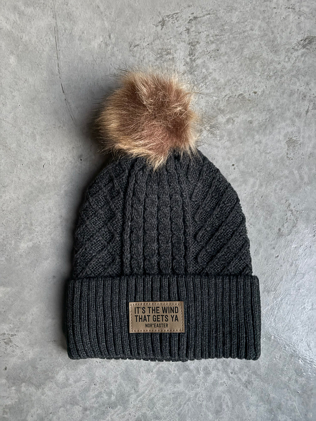 Dark gray knit beanie with a brown faux fur pom-pom on a gray concrete background