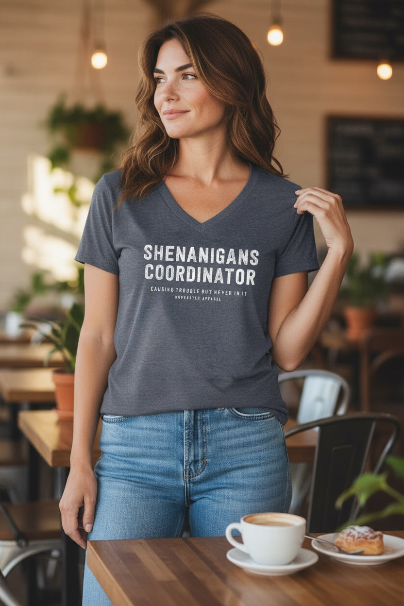 Women's vneck t-shirt that says Shenanigans coordinator on the front in white in. Set in a cafe environment. 