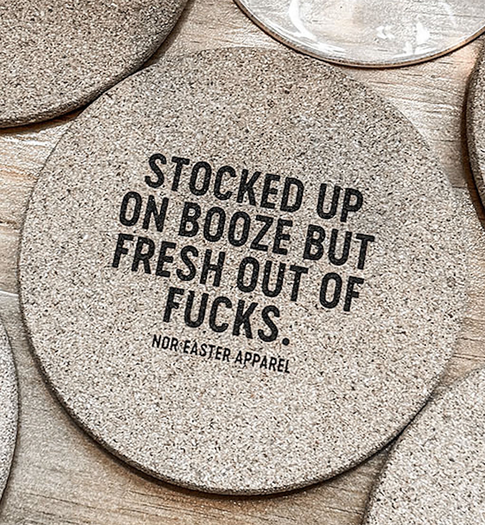 Round cork coaster with text 'Stocked up on booze but fresh out of fucks.' by Nor'easter Apparel on a wooden surface.