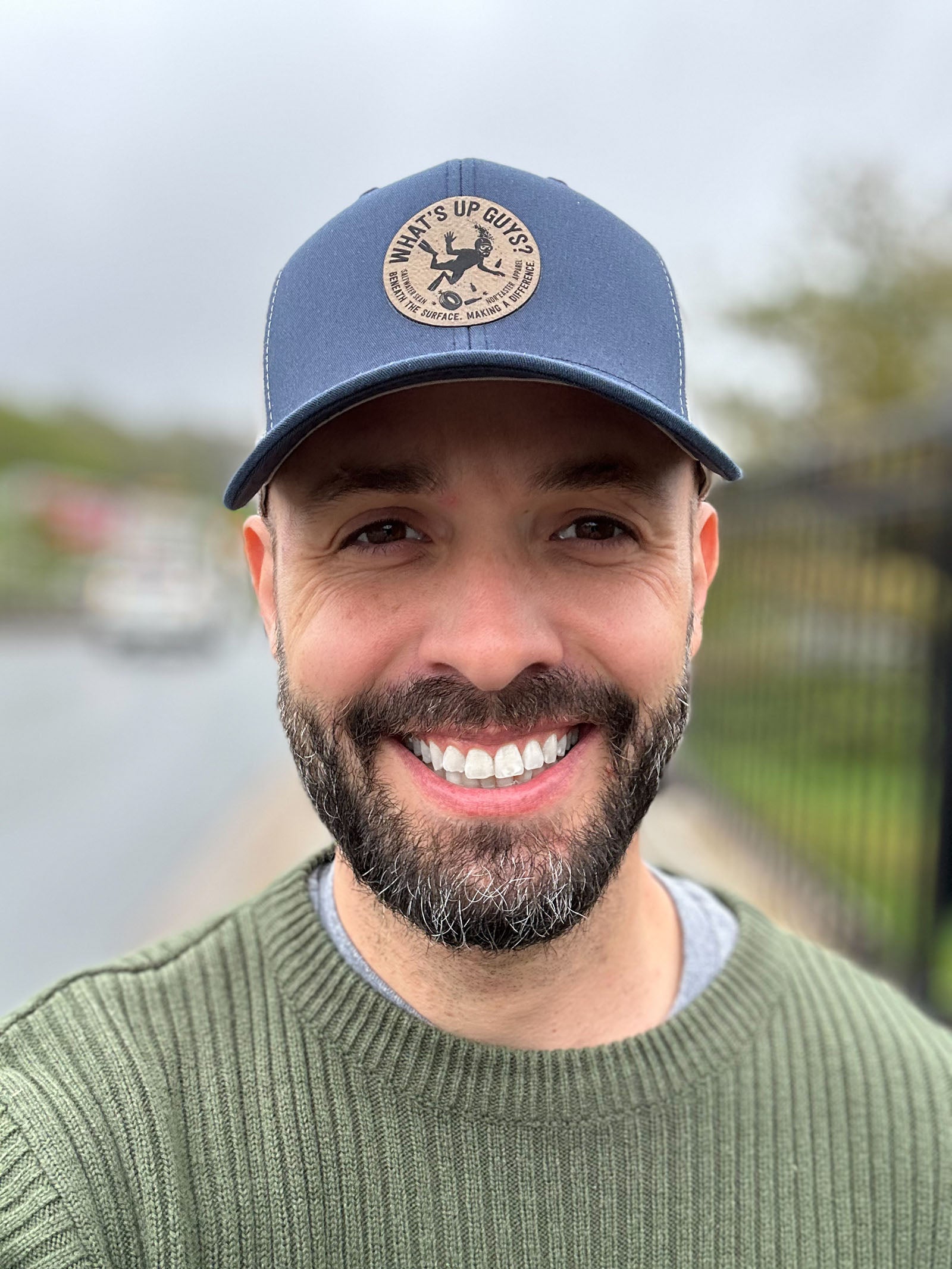 Man wearing a blue cap with a logo, smiling outdoors near water.