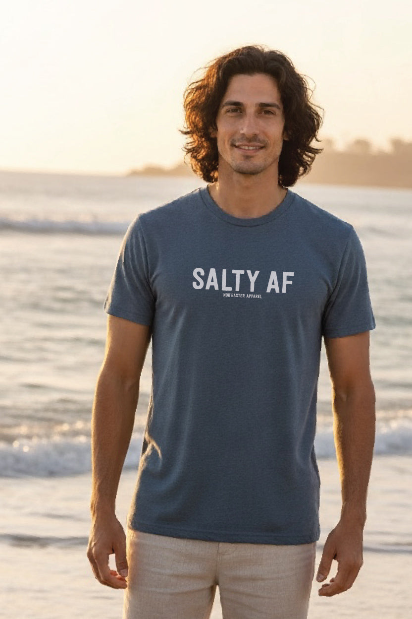 A man with long wavy hair stands on a beach wearing a heathered slate blue t-shirt with 'SALTY AF' printed in white across the chest, paired with light beige pants.