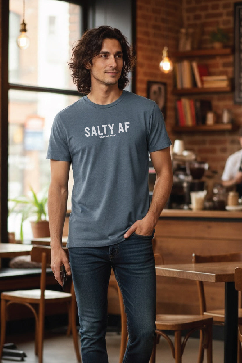 A man wearing a slate blue crewneck t-shirt with 'SALTY AF' printed in white across the chest, standing in a cozy, warm-toned coffee shop.