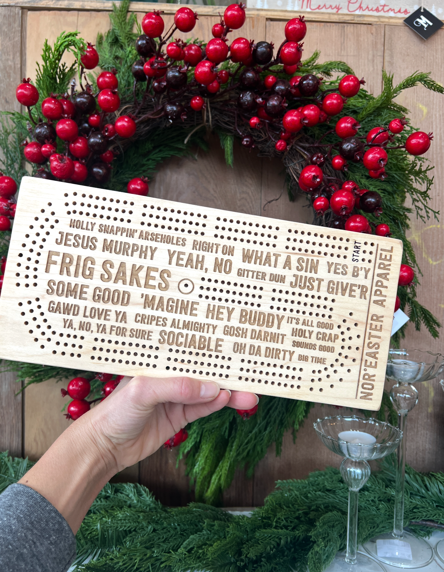 Hand holding a wooden cribbage board with text in front of a Christmas wreath.
