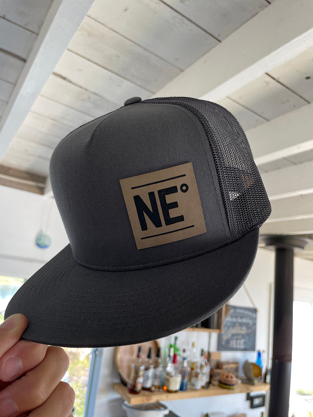 A hand holding a charcoal gray trucker hat with an adjustable back, featuring a light brown faux-suede NE° patch on the front.